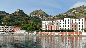 A coastal town with a prominent white building with red accents, a beach with umbrellas, and a hill with buildings in the background. - Hotel Lido Mediterranee (Taormina)