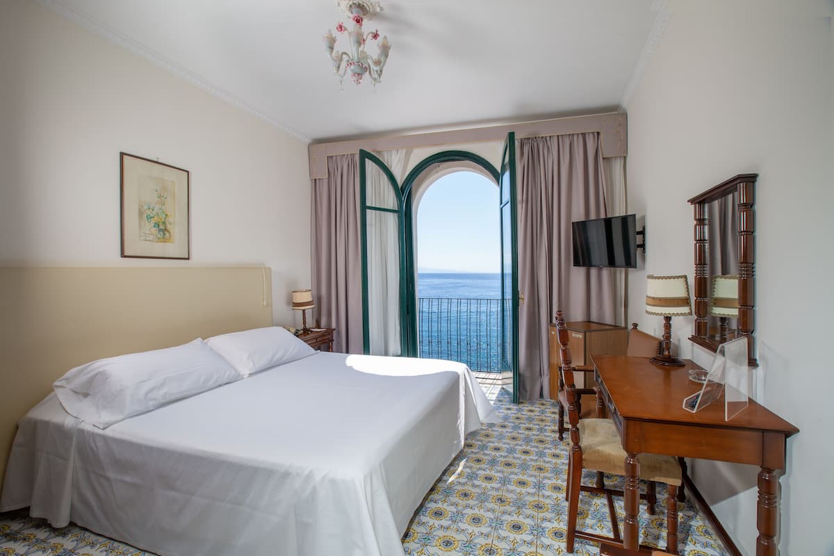 Habitación de hotel con cama, escritorio, silla, televisión y vista al mar a través de una puerta abierta.