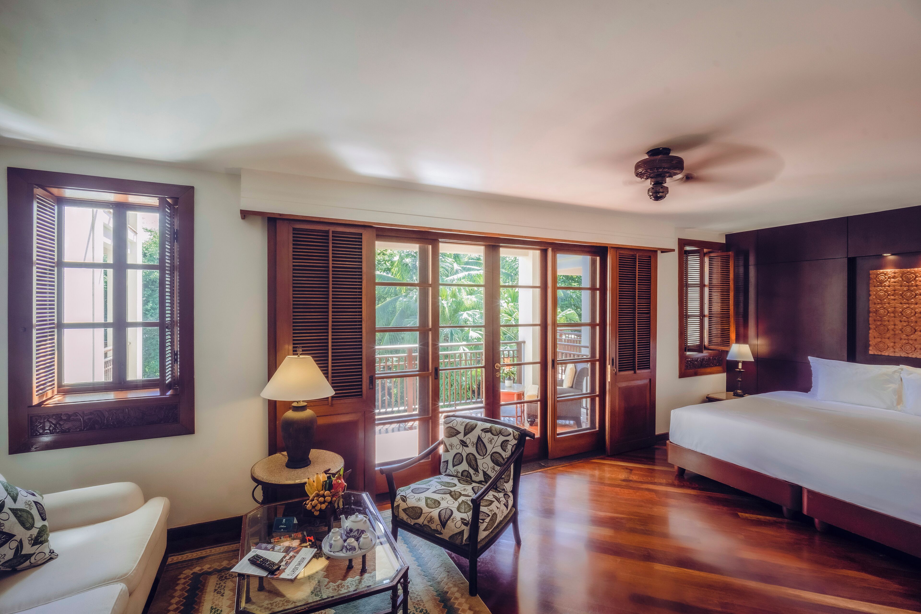 A hotel room with a bed, a chair, a table with a lamp, and large windows with shutters.