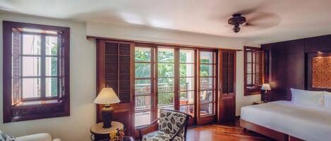 A hotel room with a bed, a chair, a table with a lamp, and large windows with shutters.
