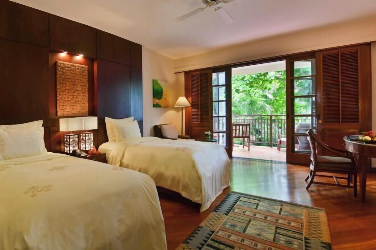 A hotel room with two beds, a balcony, a dining table, and a view of greenery.