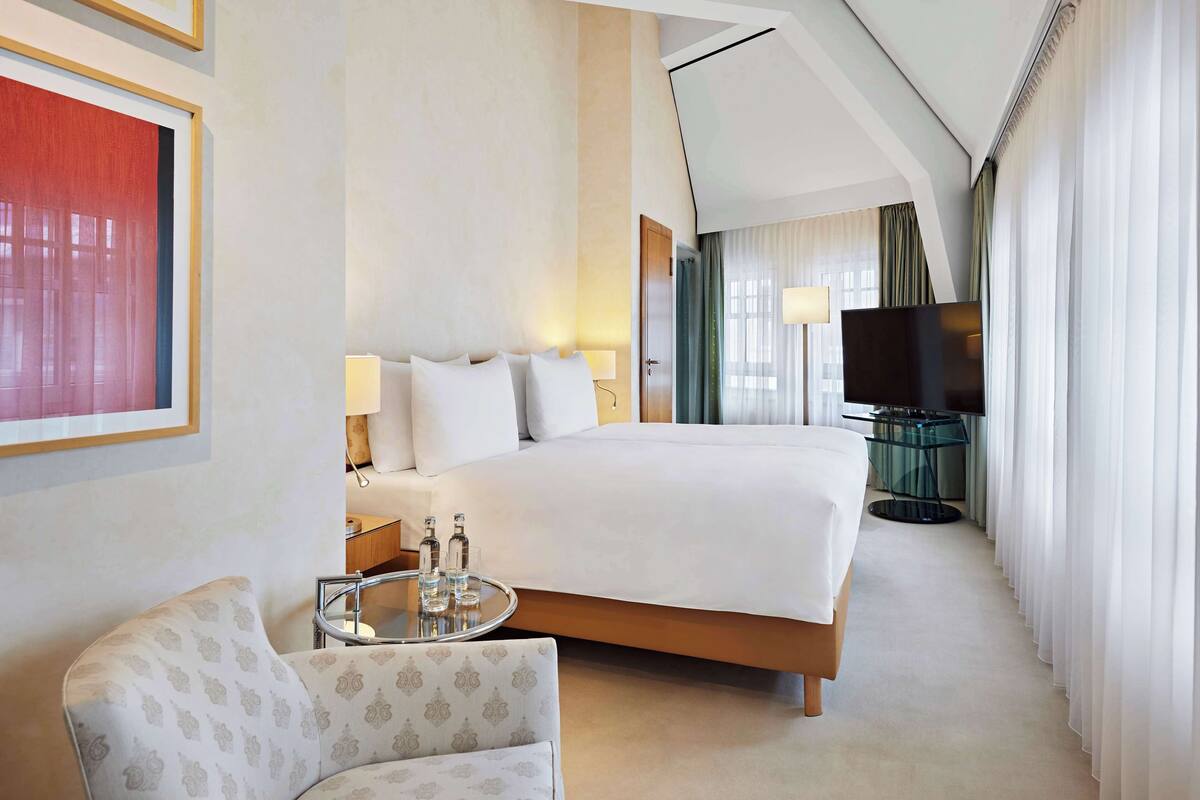 A hotel room with a large bed, a TV, a glass table with bottles, and a chair.