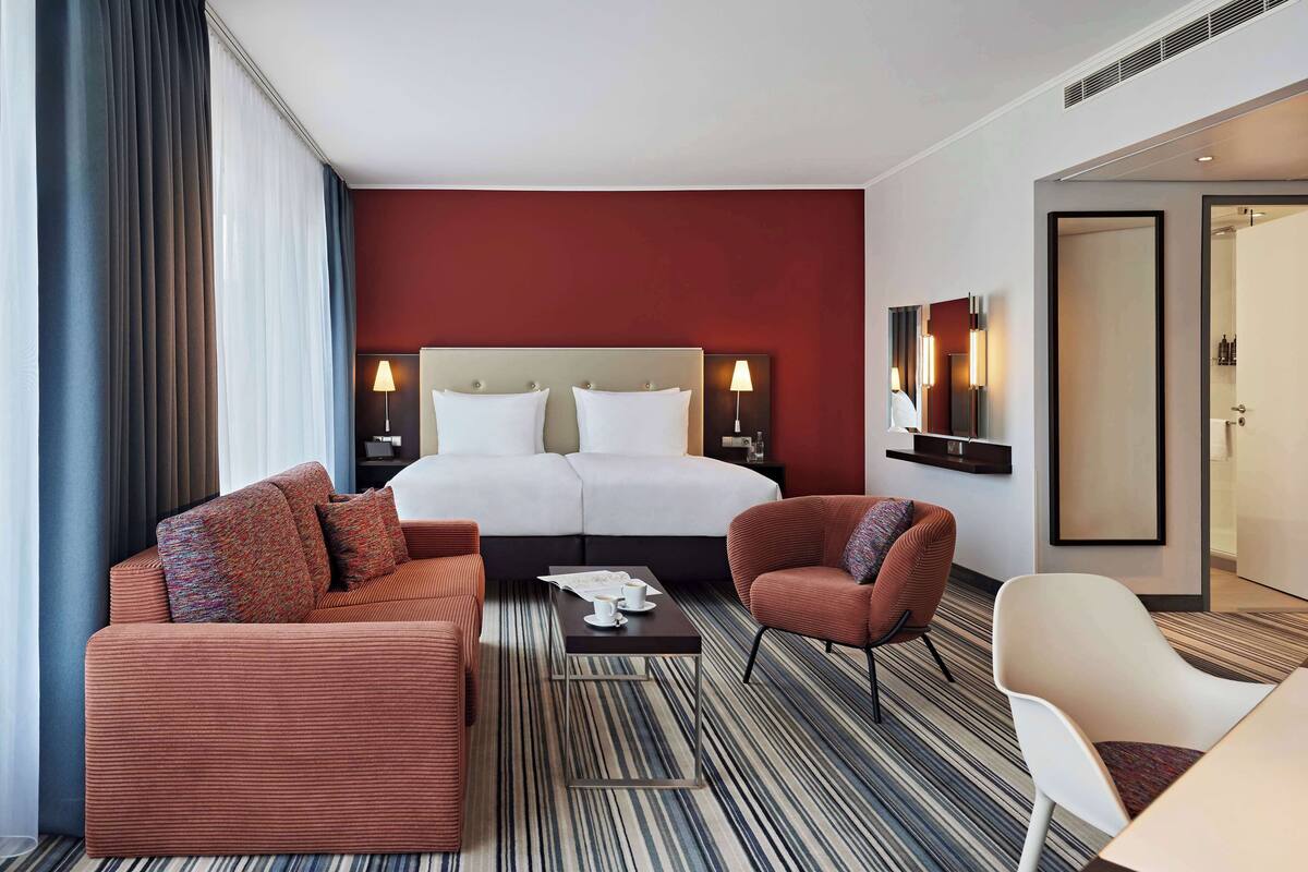 A hotel room with a large bed, two bedside tables with lamps, a sofa, a chair, and a striped carpet.