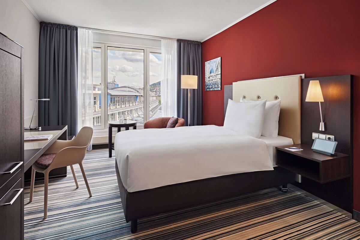 A hotel room with a large bed, a desk, a chair, and a view of the city through the window.