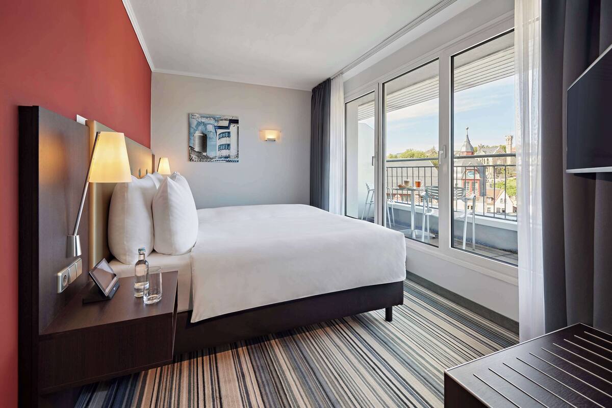 A hotel room with a large bed, a bedside table with a lamp, a glass of water, and a painting on the wall.