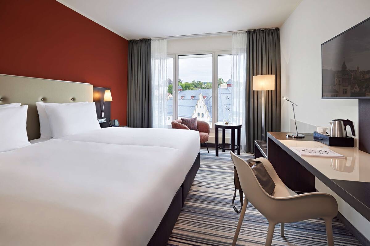 A hotel room with a large bed, a desk, a chair, a small table, and a view of buildings through the window.
