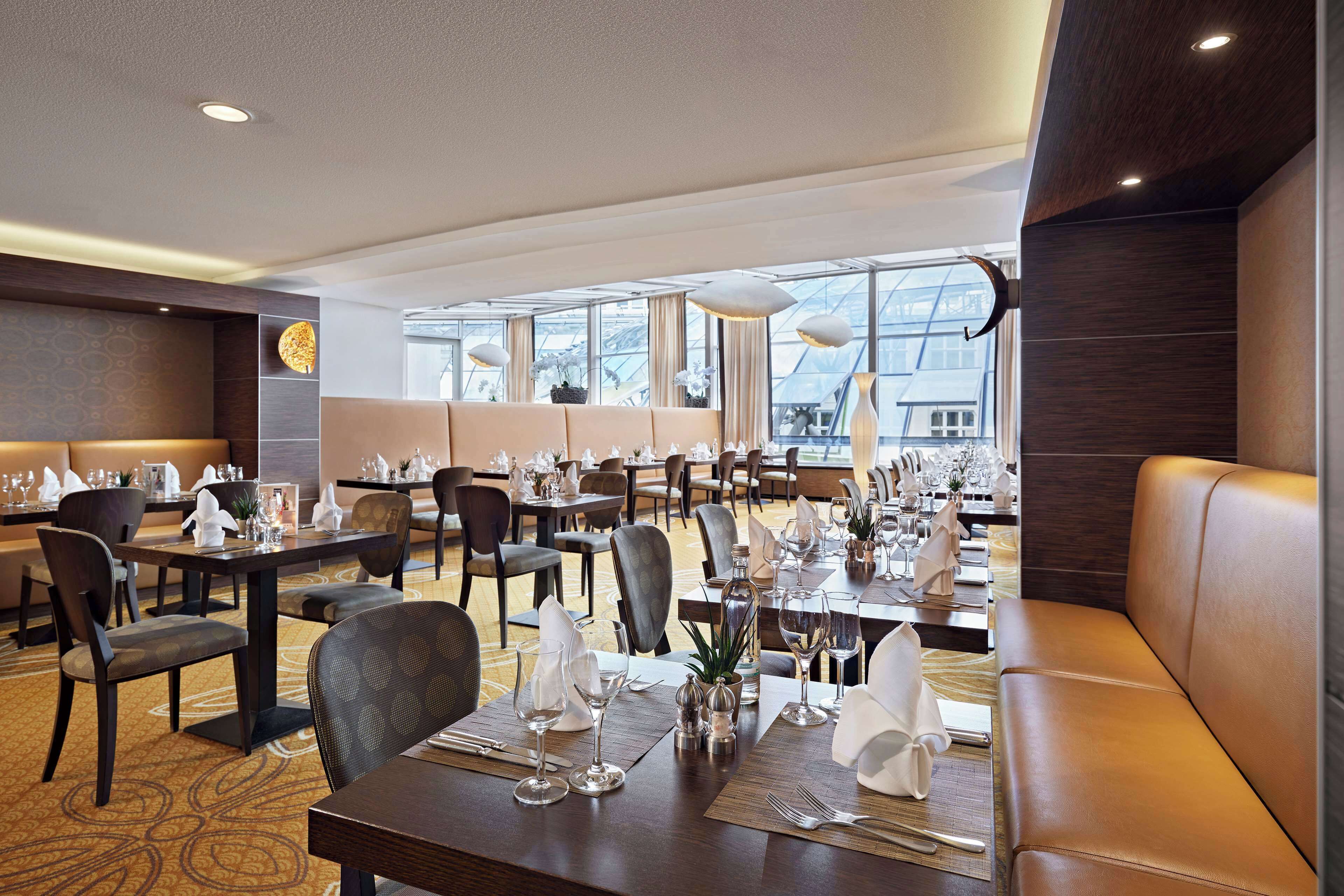 A modern restaurant with multiple dining tables, cushioned seating, and large windows offering views of the city.