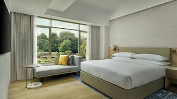 A modern hotel room with a large bed, a sofa, a small table, and a view of greenery outside.