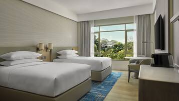 A hotel room with two beds, a desk, a chair, a TV, and a large window with a view of greenery.