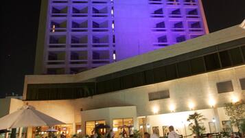 Ein Hotel mit Außenbereich zum Essen und einem Pool, das sich durch modernes Design, einen markanten Turm und eine beleuchtete Fassade auszeichnet.