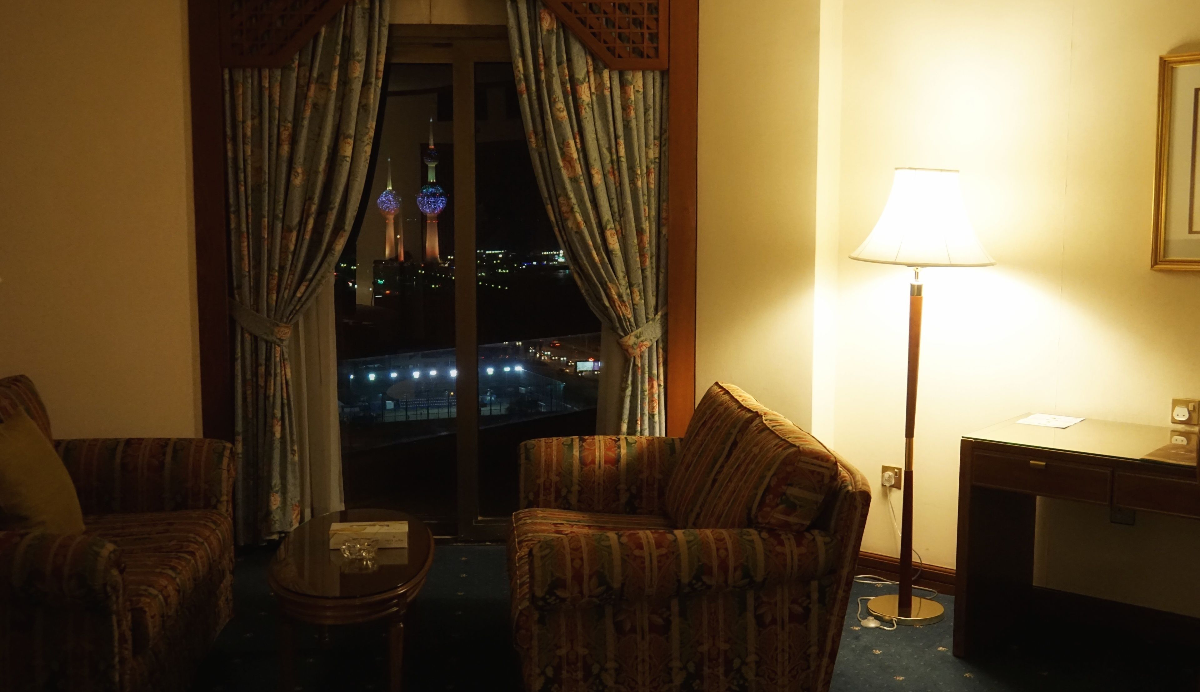 Ein Hotelzimmer mit zwei Sesseln, einem kleinen Tisch, einer Lampe und Blick auf eine nächtliche Stadtlandschaft.