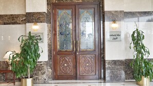 A wooden double door with intricate carvings, flanked by two wall-mounted light fixtures and two potted plants. - Safir International Hotel Kuwait (Kuwait City)