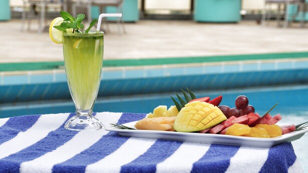A glass of green beverage with a straw and mint garnish, a plate of assorted fruits, and a poolside setting with a blue and white striped towel. - Safir International Hotel Kuwait (Kuwait City)