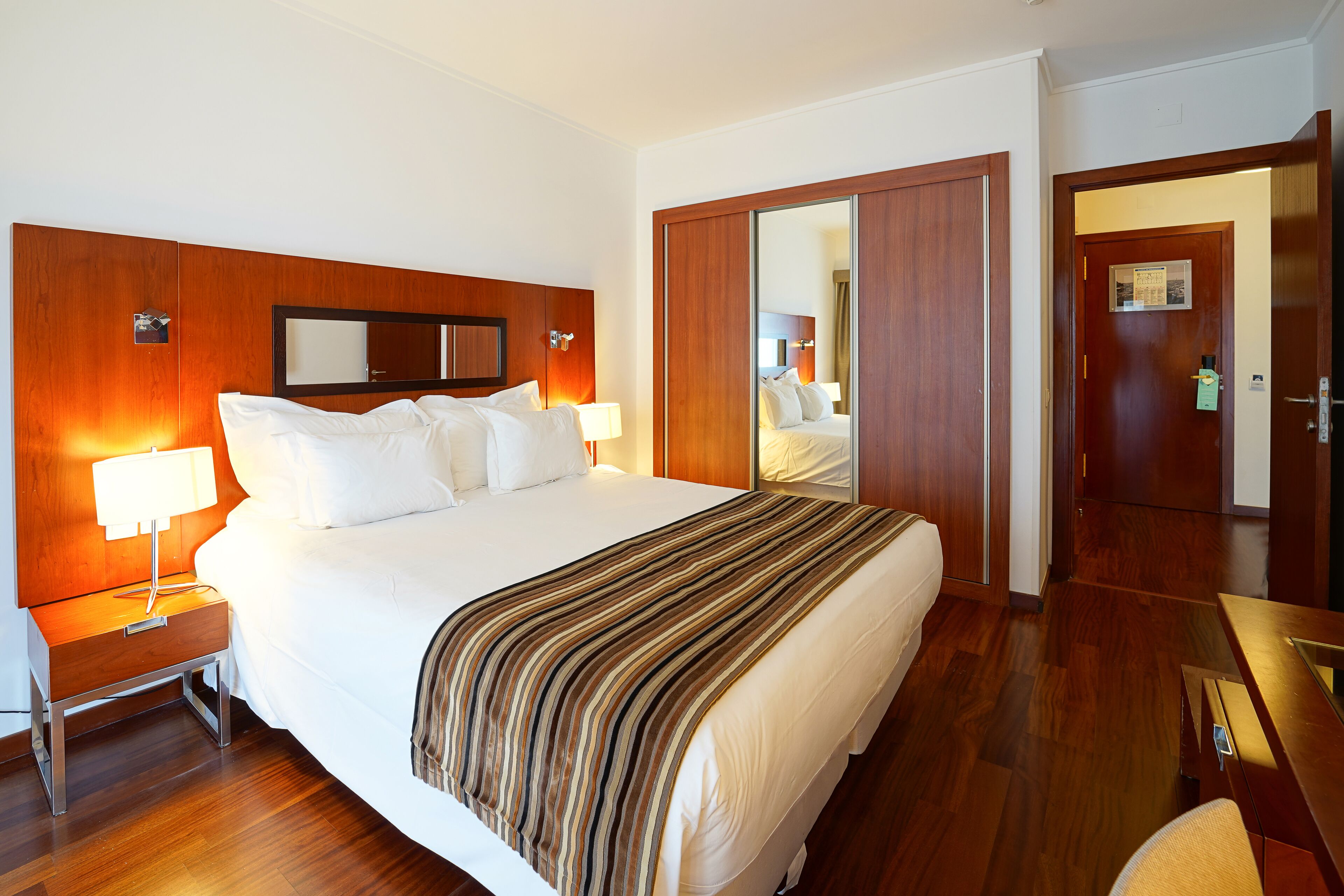 Ein Hotelzimmer mit einem großen Bett, einem Holz-Kopfteil, einem Nachttisch mit Lampe, einem Spiegel und einem Kleiderschrank.