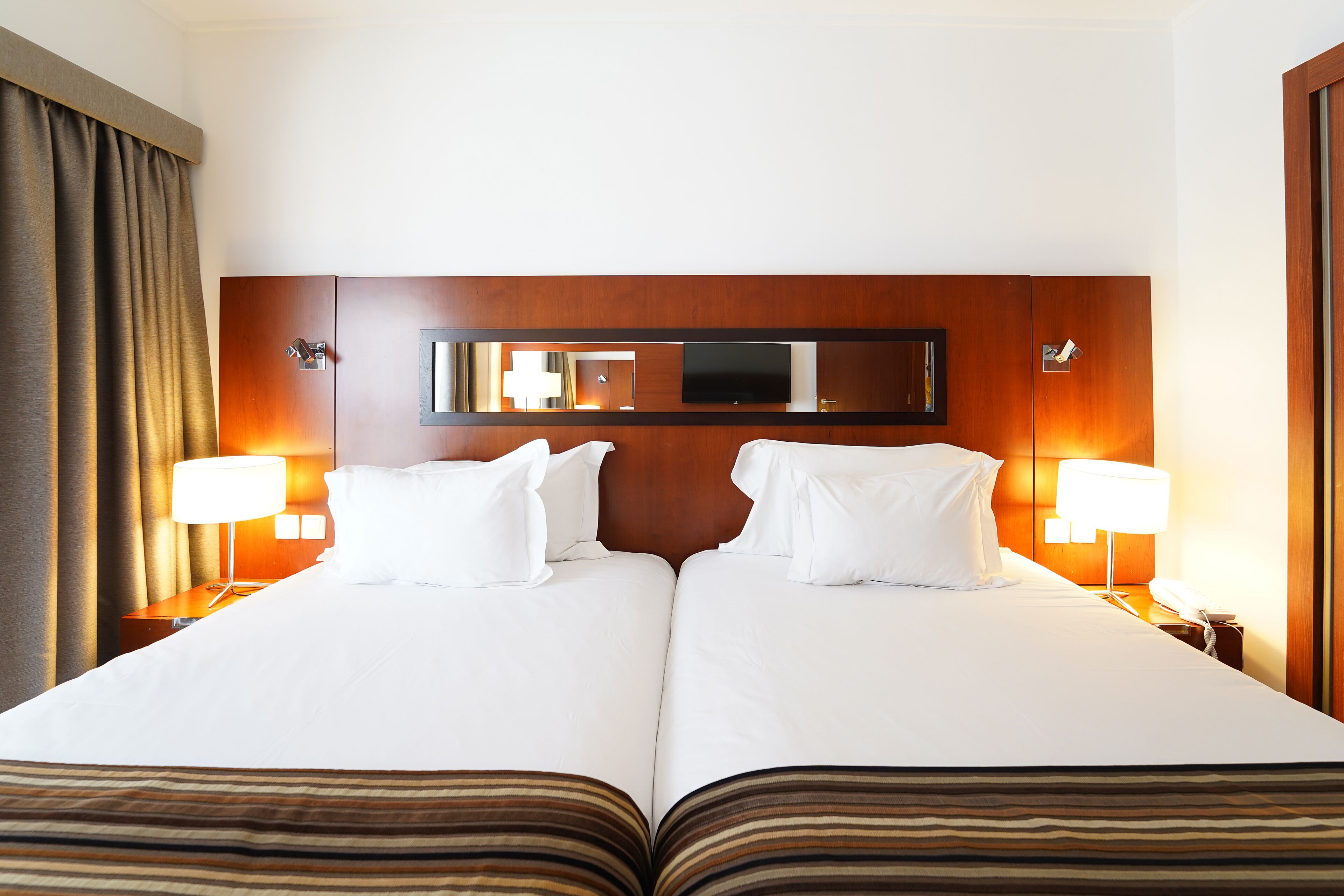 A double bed with white linens, a wooden headboard, a flat-screen TV mounted above the bed, two bedside lamps, and a large mirror.