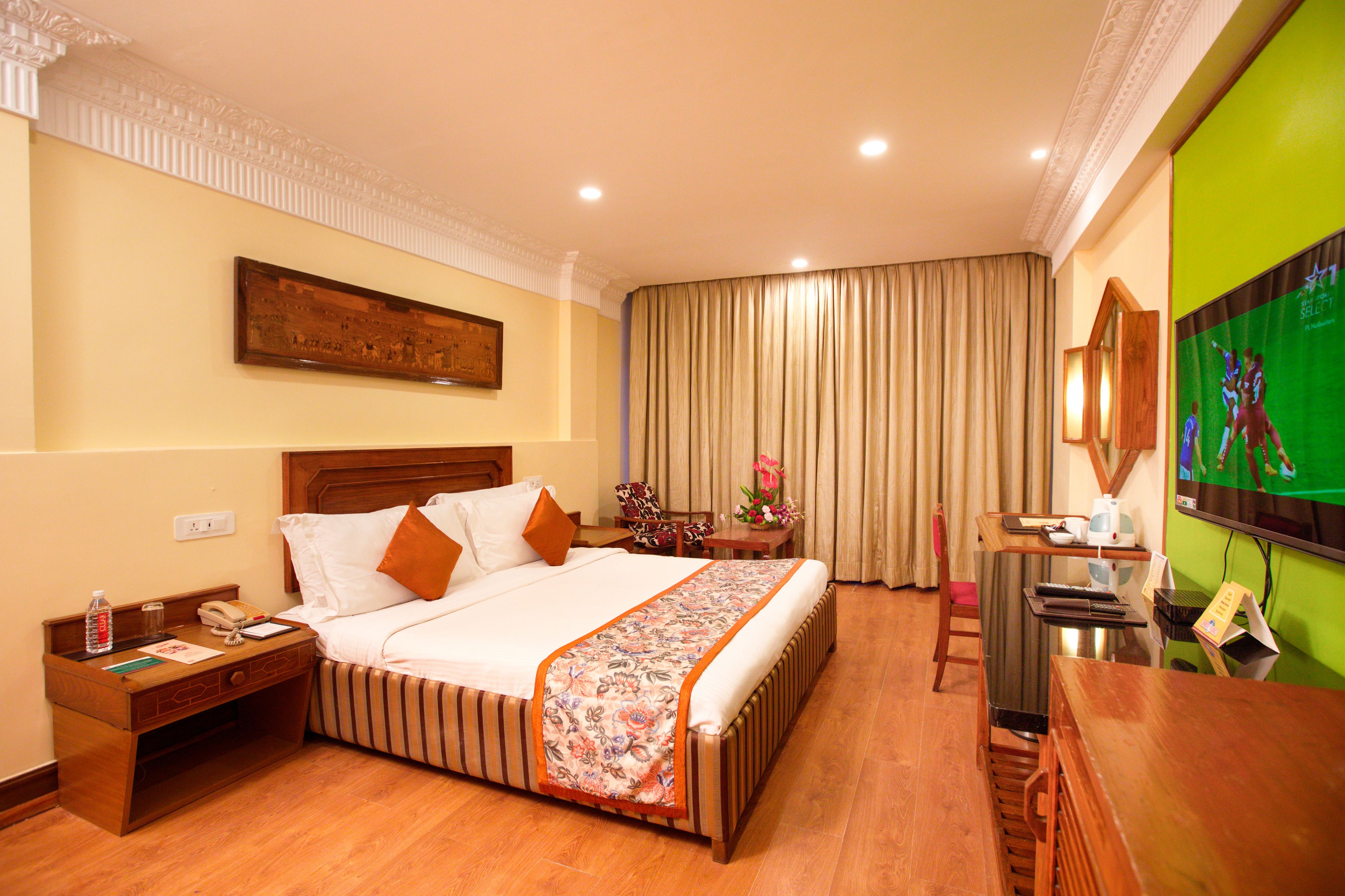 A hotel room with a large bed, a wooden nightstand, a desk, a chair, and a television.
