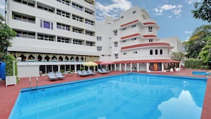A swimming pool with sun loungers and umbrellas, surrounded by a multi-story building with balconies and a garden area. - Ambassador Pallava (Chennai)