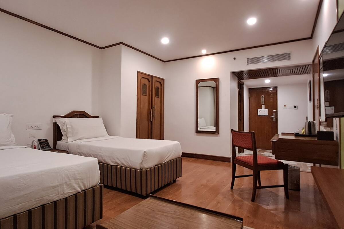 A hotel room with two beds, a wooden floor, a chair, a desk, a mirror, and a TV.