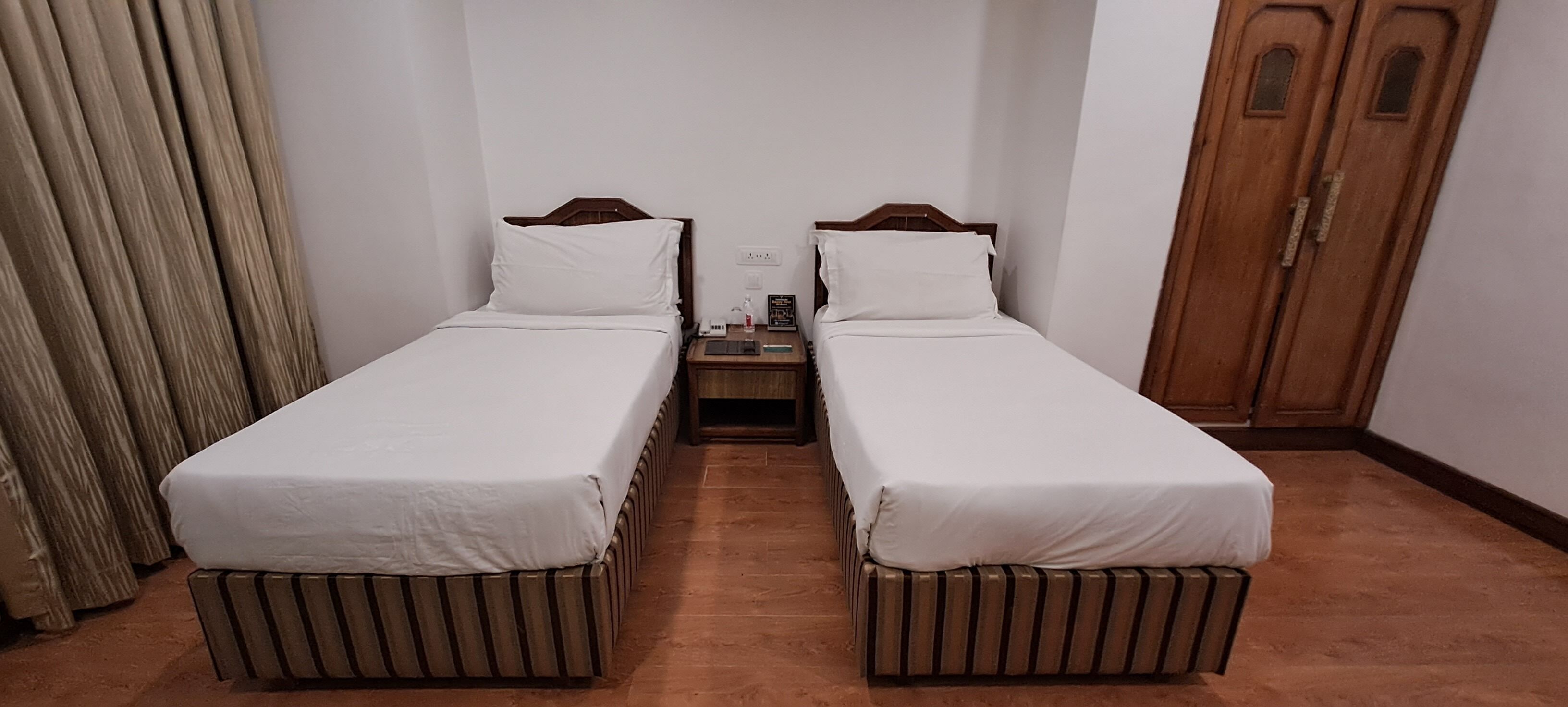 A room with two single beds, wooden headboards, a nightstand with a lamp, and a wooden wardrobe.