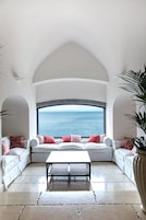 Una habitación con un gran ventanal arqueado con vistas al mar, un sofá blanco con cojines rojos y una pequeña mesa en el centro.