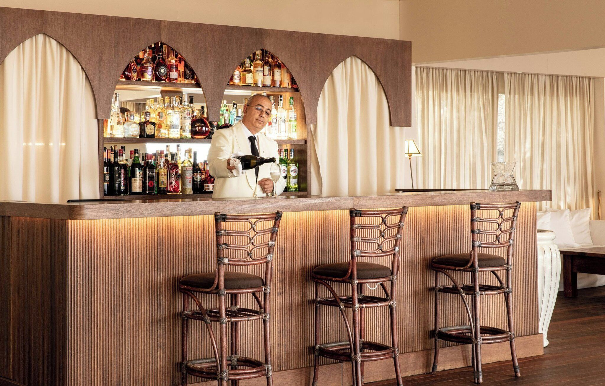A bar area with a bartender, a selection of liquor bottles, and three wicker bar stools.