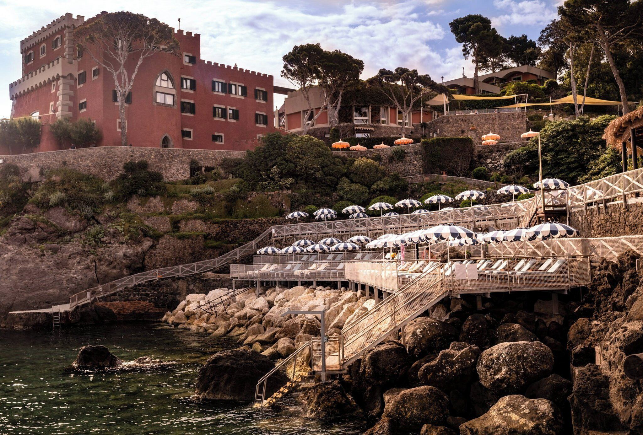 A coastal hotel with a castle-like building, outdoor seating with umbrellas, and a rocky shoreline.