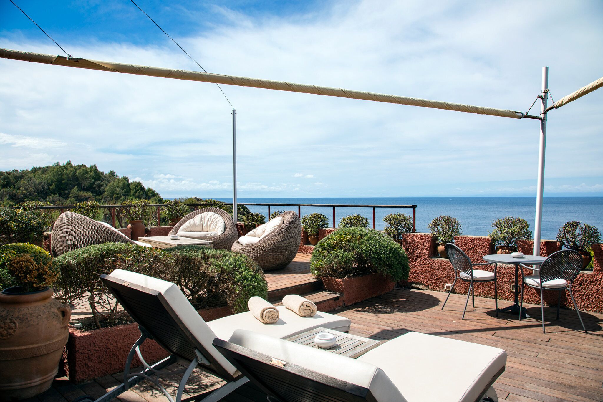 A sunlit outdoor seating area with lounge chairs, a table, and potted plants, overlooking a body of water.