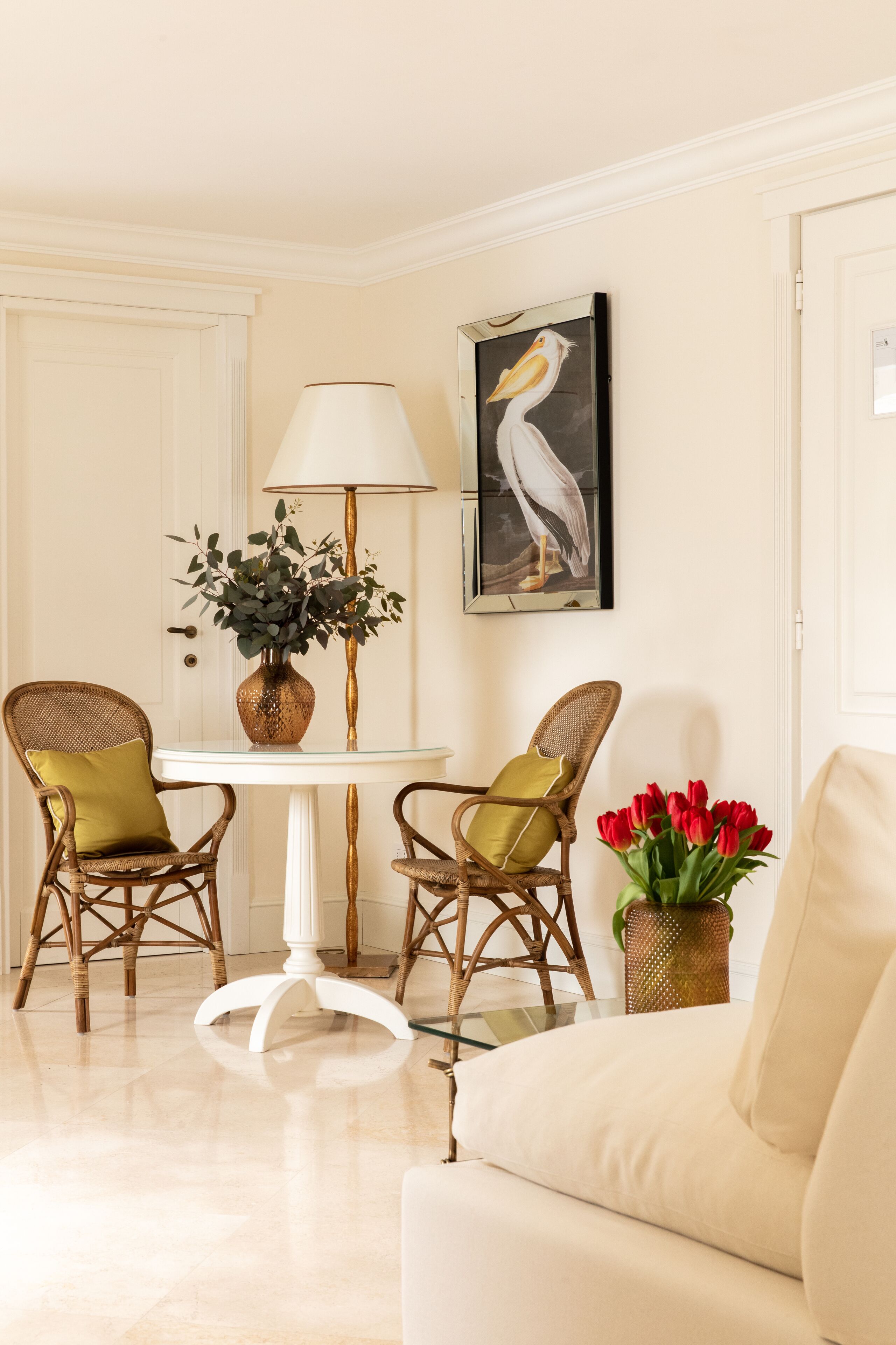 Una habitación con un sofá blanco, dos sillones de mimbre con cojines, una mesita redonda con una lámpara, un florero con flores rojas y un cuadro enmarcado en la pared.