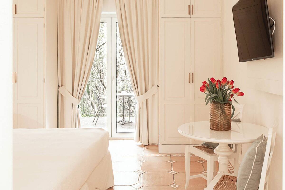 Una habitación con una cama, una mesa redonda con un jarrón de flores rojas, y vista al exterior a través de un ventanal.