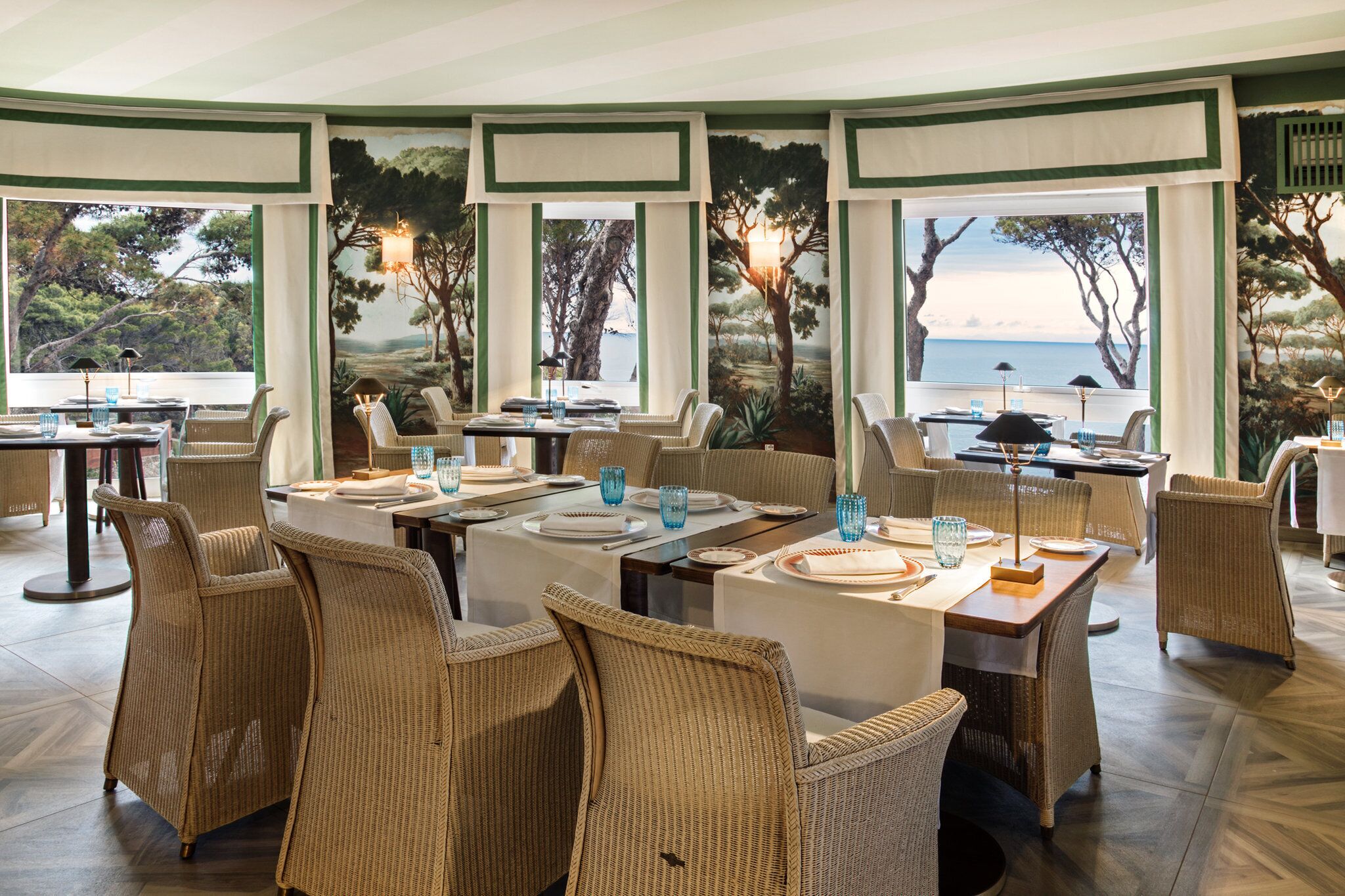 Een restaurant met rieten meubels, gedekte tafels met borden en glazen, en grote ramen met uitzicht op bomen en de zee.