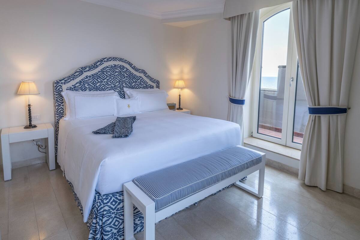 Une chambre d’hôtel bien aménagée, avec un grand lit, des lampes de chevet, un banc et une vue sur la mer par la fenêtre.