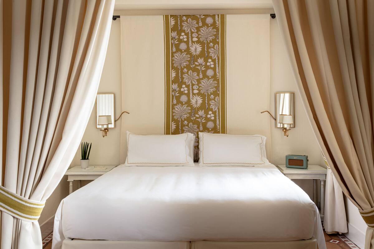 Une chambre d’hôtel avec un grand lit, deux tables de chevet, une petite table, une plante en pot et un panneau décoratif mural.