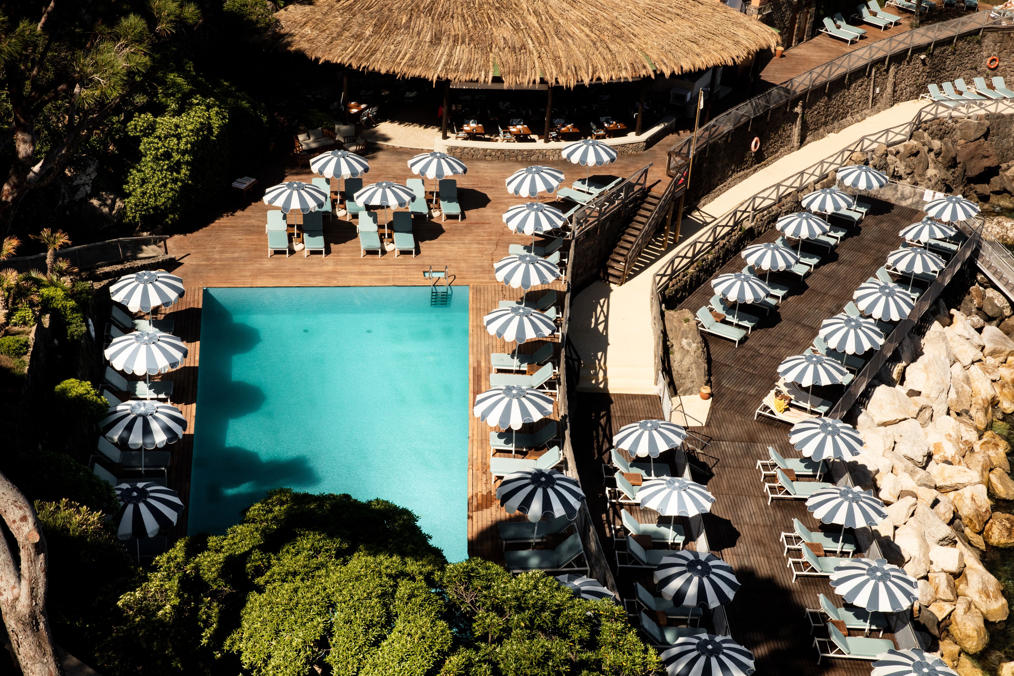 Vista aérea de un complejo turístico con una piscina central rodeada de sillones de playa y sombrillas, una estructura con techo de paja y una costa rocosa.