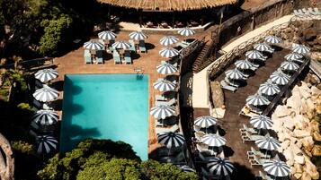 Vista aérea de un complejo turístico con una piscina central rodeada de sillones de playa y sombrillas, una estructura con techo de paja y una costa rocosa.