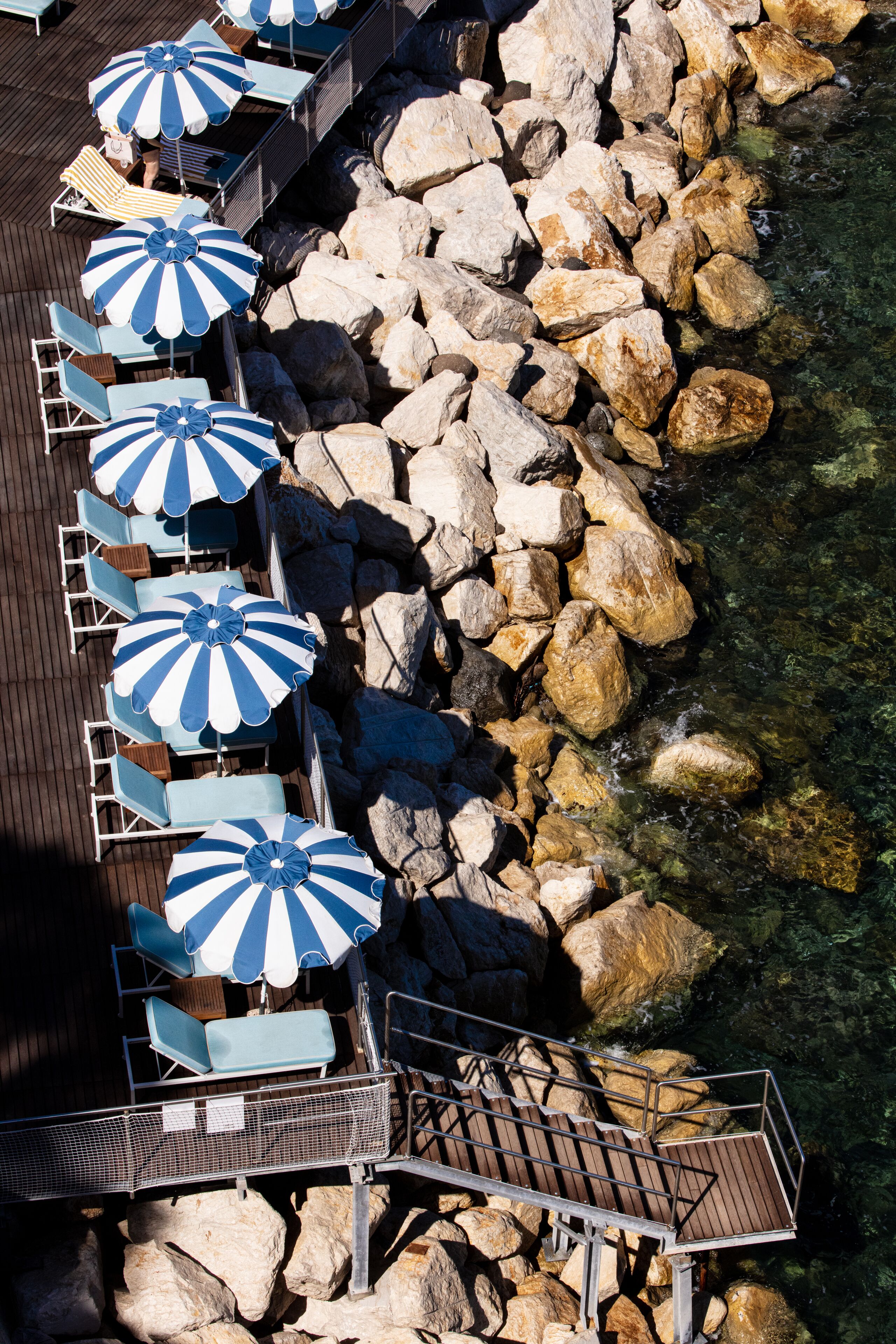 A deck with lounge chairs and blue and white striped umbrellas overlooking a rocky shoreline.