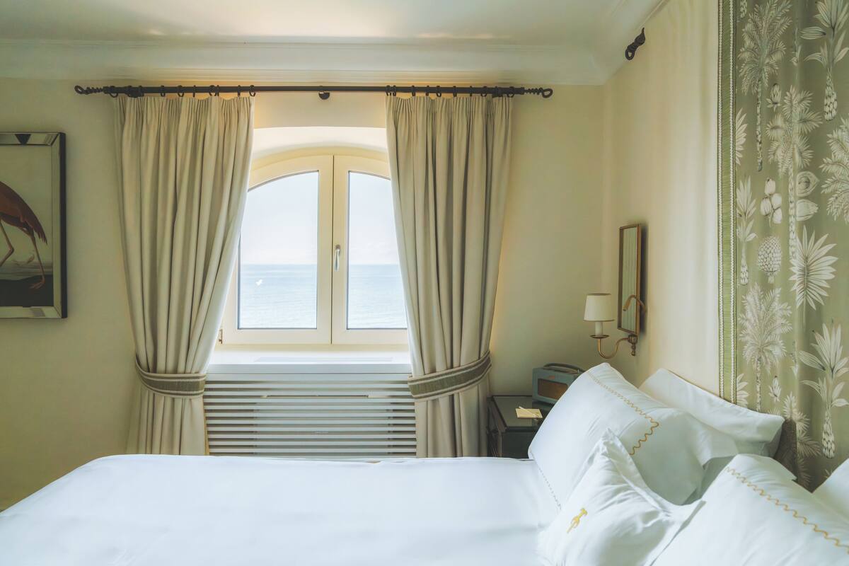 Une chambre d’hôtel avec un lit, des rideaux, une fenêtre donnant sur un paysage agréable, une table de chevet et une lampe.