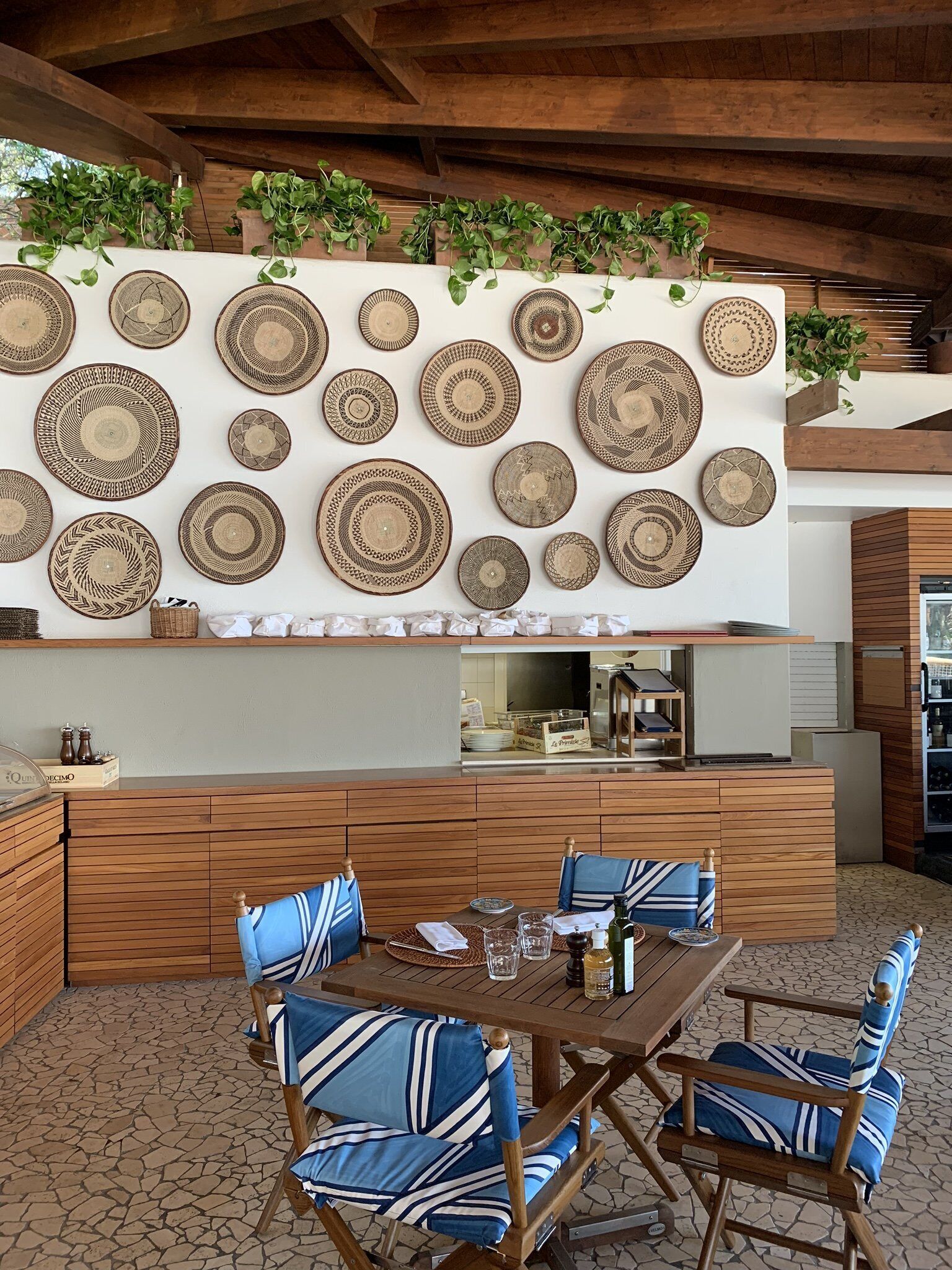 Un espace extérieur aménagé pour les repas, avec une table et des chaises en bois, entouré d’un mur décoré d’assiettes tressées.