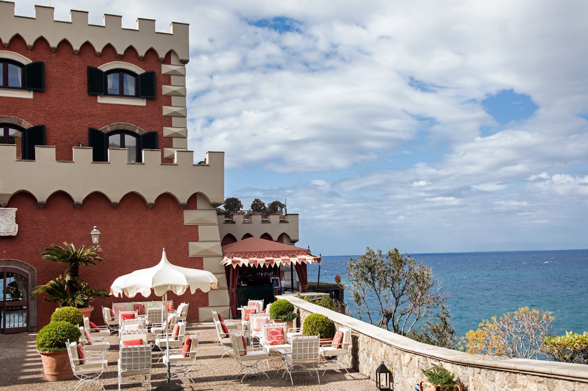 A seaside hotel with a castle-like facade, outdoor seating with umbrellas, and a view of the sea.