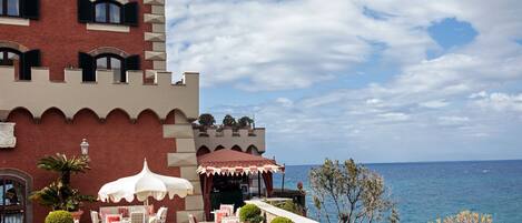 A seaside hotel with a castle-like facade, outdoor seating with umbrellas, and a view of the sea.