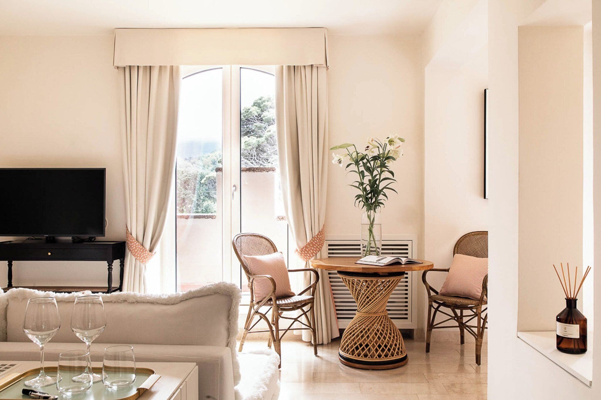 Een moderne woonkamer met een flatscreen televisie, een bank, twee rieten stoelen, een ronde tafel met een bloevas en een raam met gordijnen.