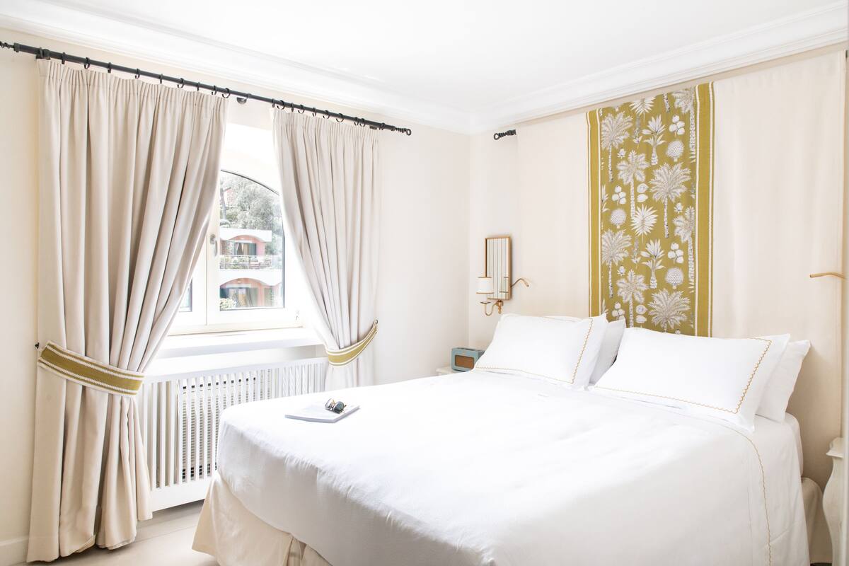 Un lit bien fait, avec du linge de lit blanc, une fenêtre ornée de rideaux beiges et un mur décoré d’un motif floral.