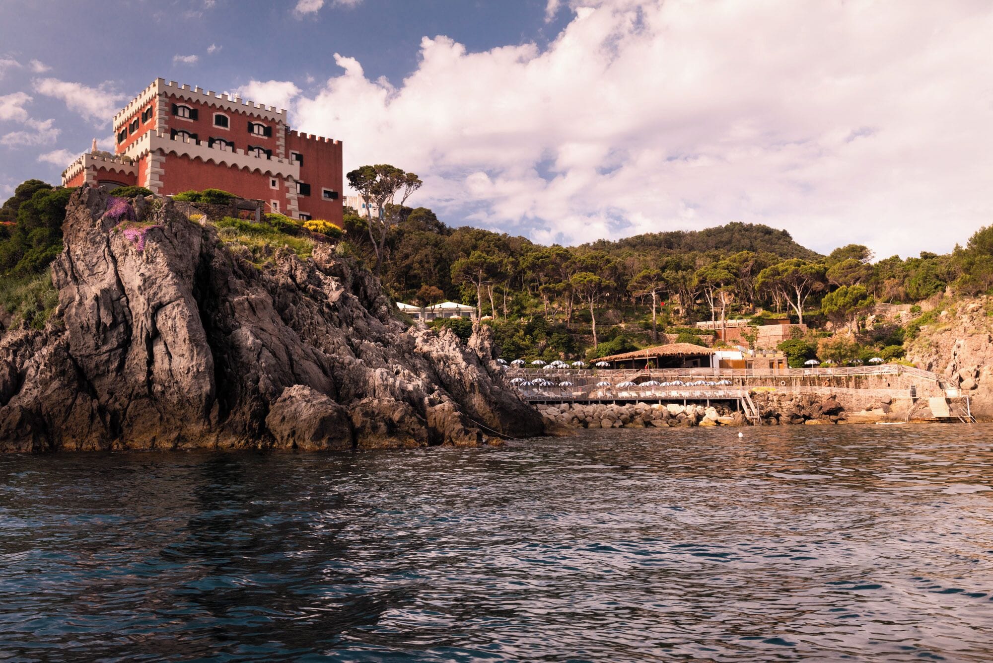 A castle-like building on a rocky cliff by the sea, with a dock and umbrellas in the background.