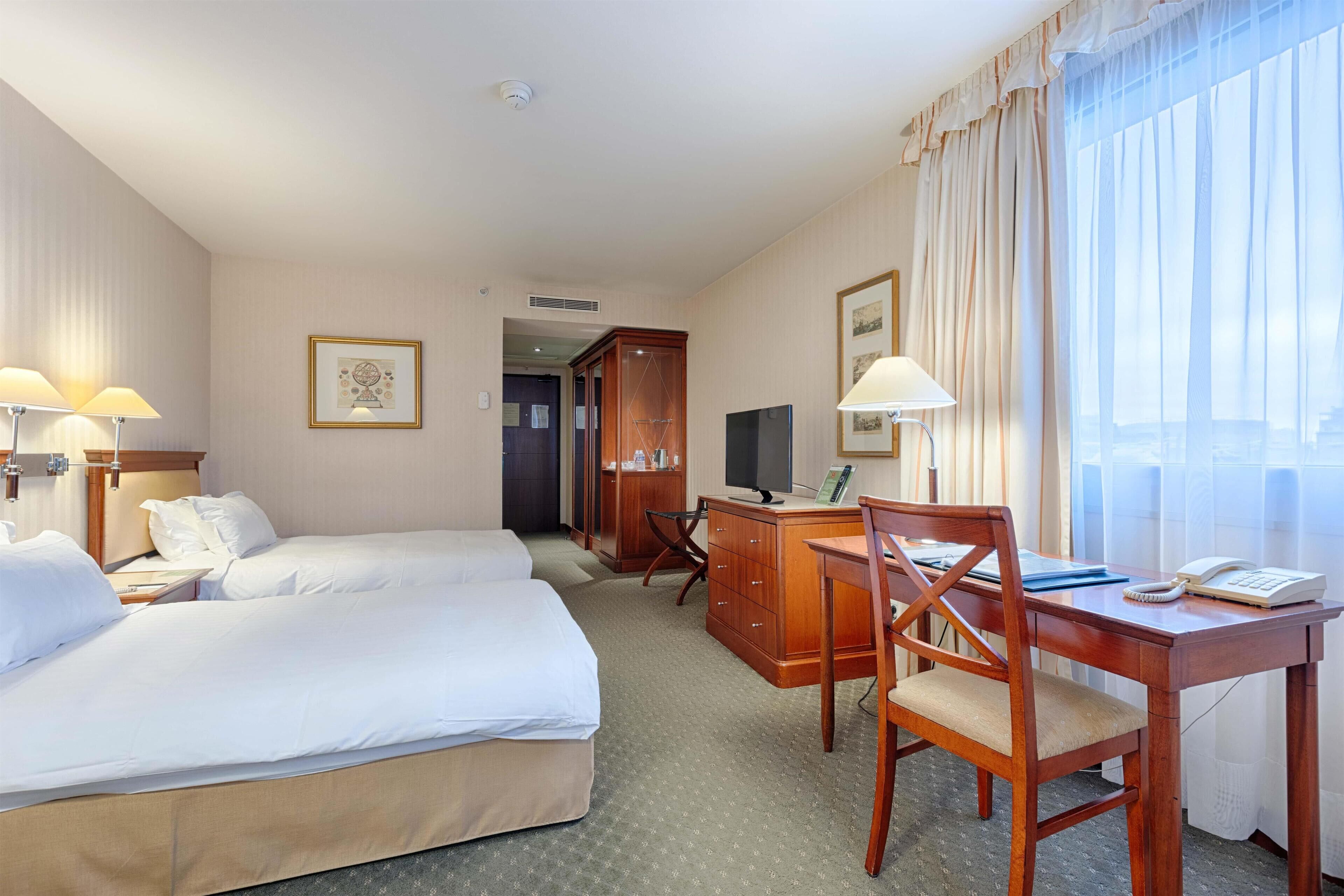 A hotel room with two beds, a desk, a chair, a television, and a window with curtains.