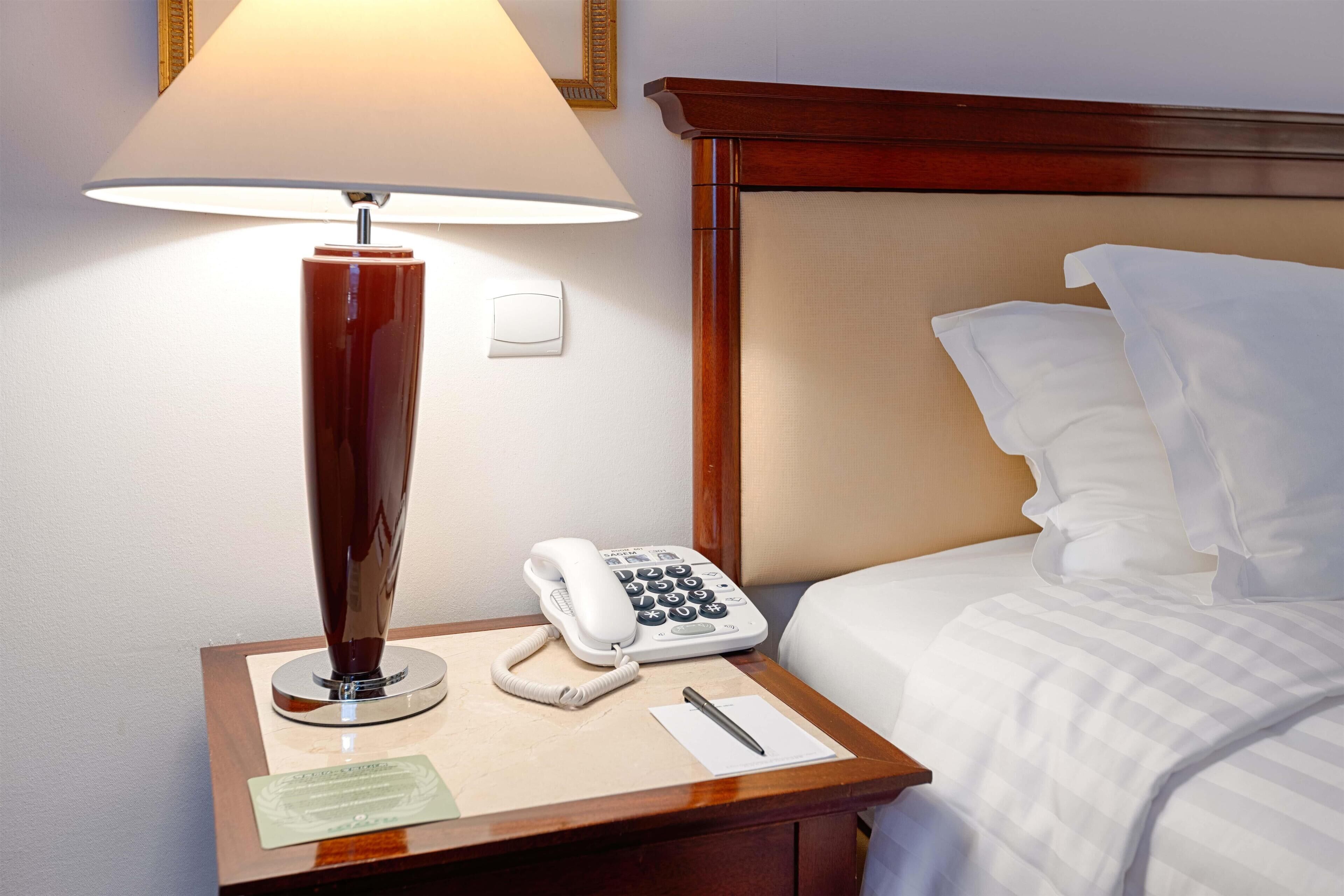 A hotel room with a bedside lamp, a telephone, a notepad, and a pen on a wooden nightstand.