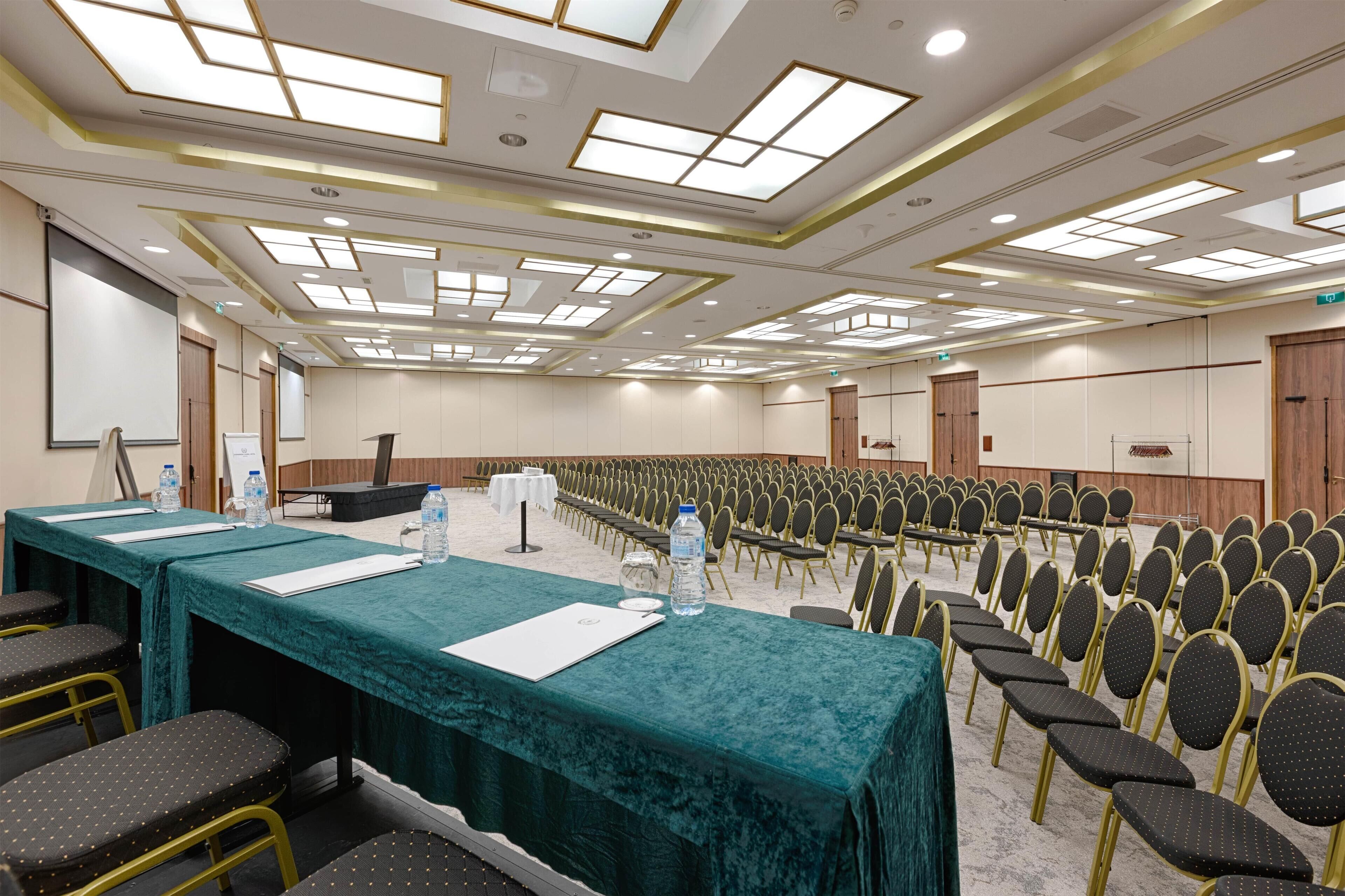 Een congrescentrum met rijen stoelen, een lange tafel met een groene tafelkleed en een podium.