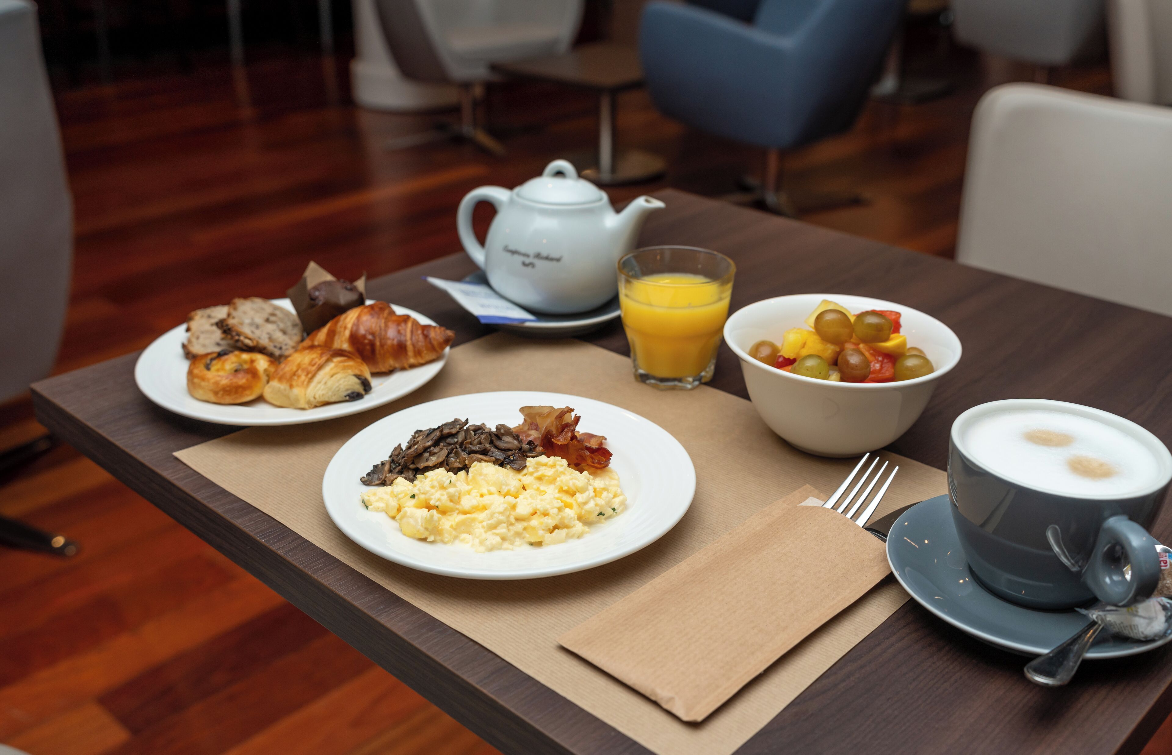 A breakfast table with croissants, scrambled eggs, bacon, and a bowl of fruit.