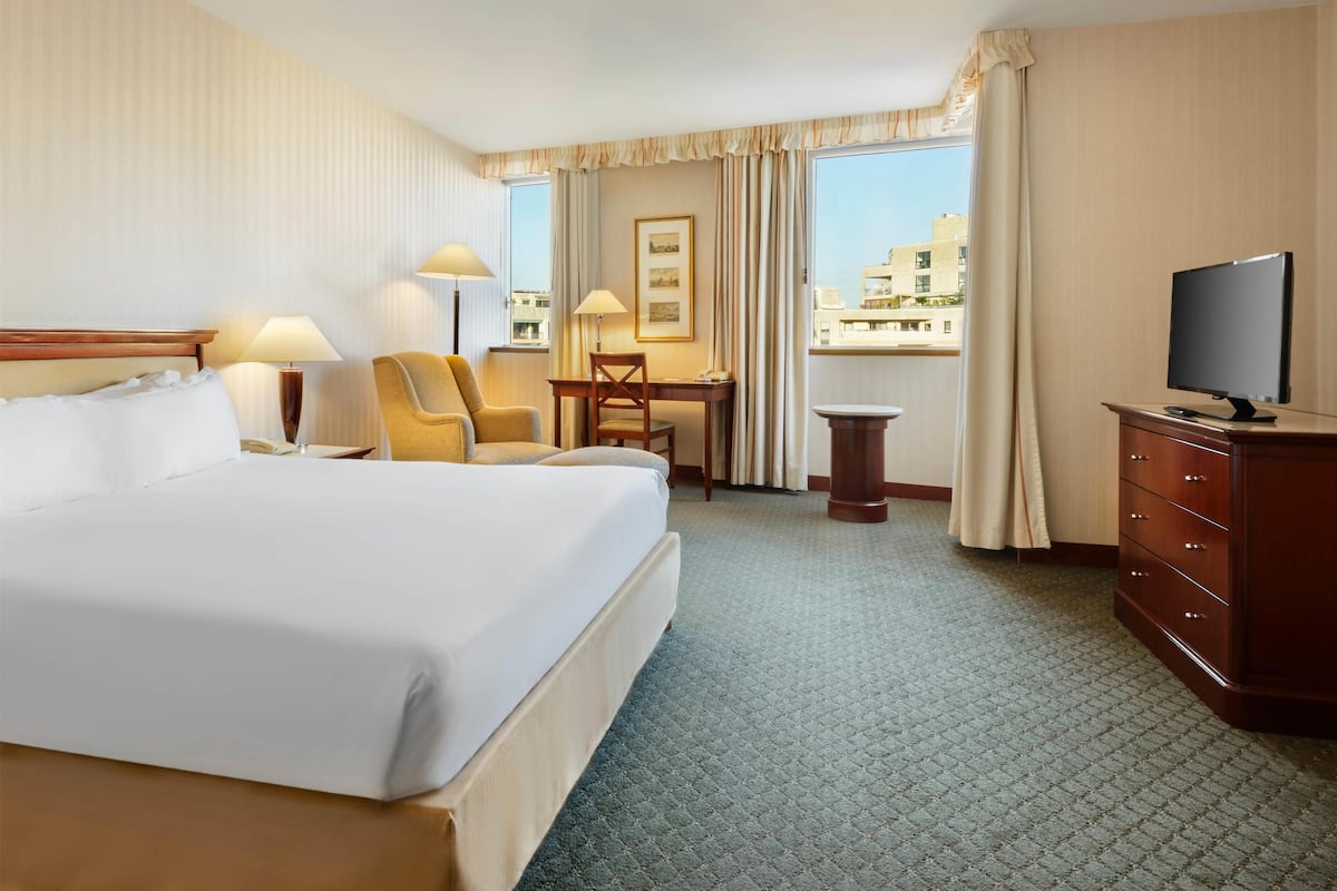 A hotel room with a large bed, a desk with a chair, a television, and a view of buildings through the window.