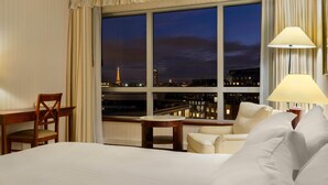 Quarto de hotel com uma janela grande, uma cama, uma escrivaninha, uma cadeira e um abajur.