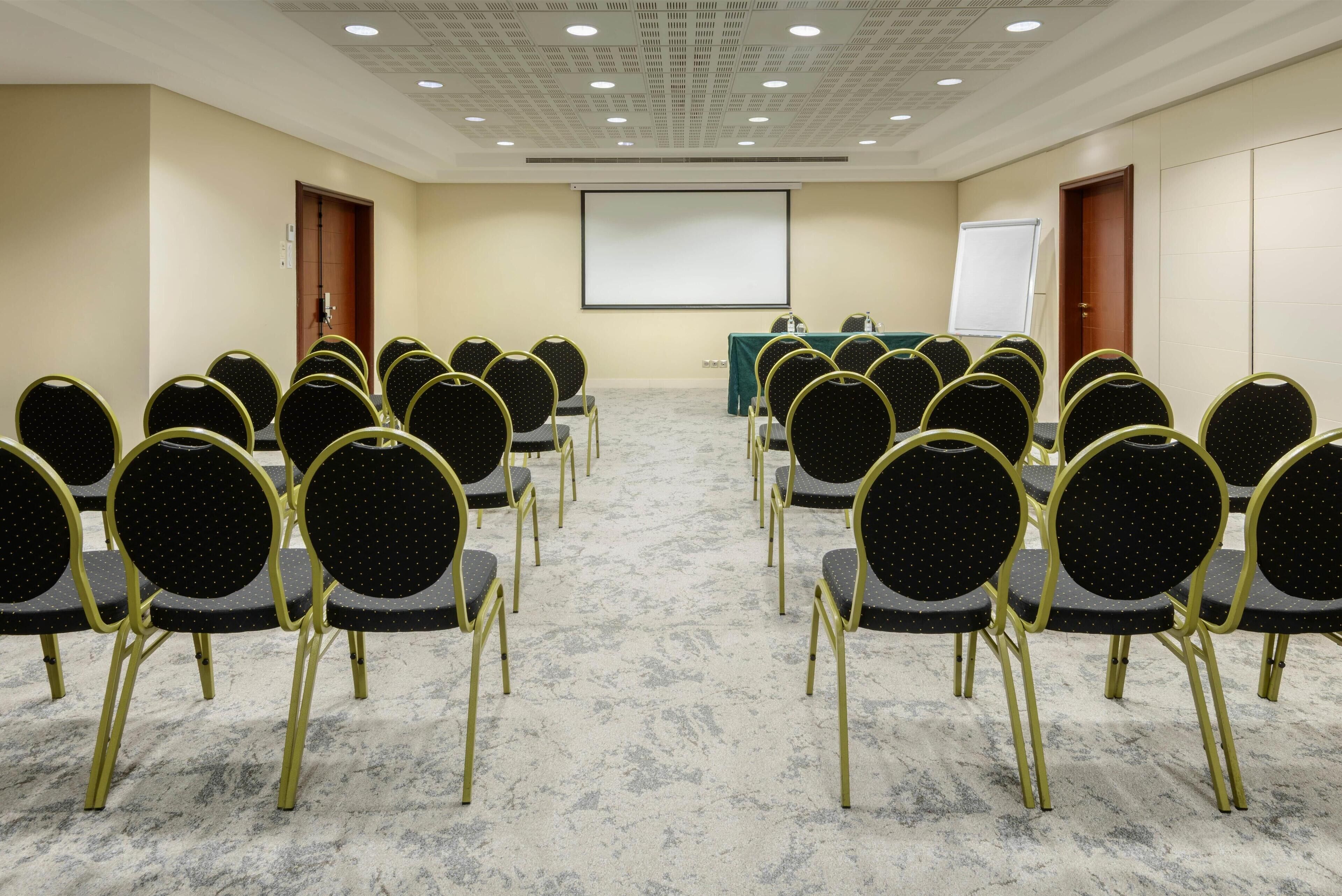 A conference room with rows of chairs facing a projector screen, a whiteboard, and a table with a green tablecloth.