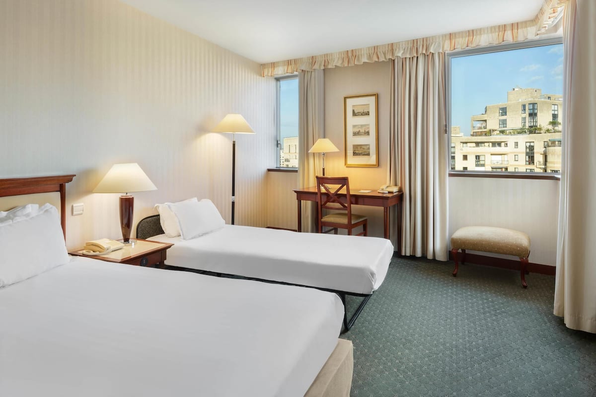 A hotel room with two beds, a desk, a chair, and a view of the city.